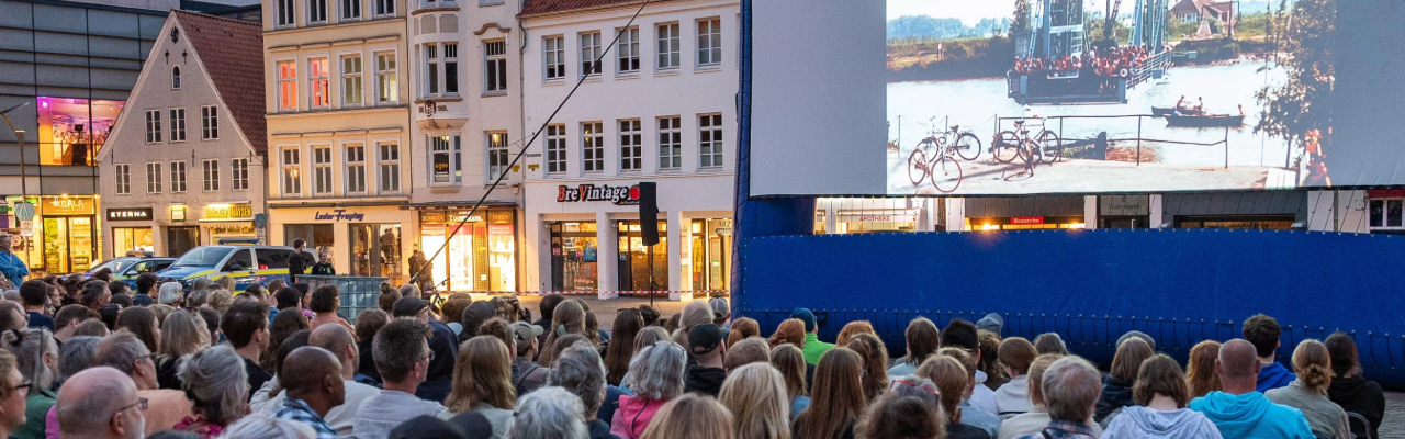 Menschen sitzen vor einer Leinwand in Reihen auf einem Platz vor Gebäuden.